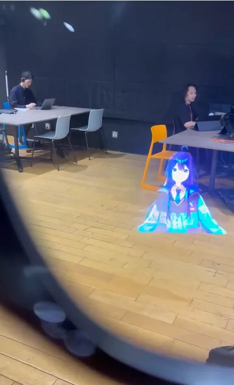 A photograph taken through the lens of a Ray-Ban Meta Display: Alice rendered standing on the wooden floor of a room, with two people working at desks behind her.